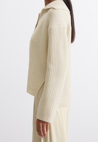 Pull tricoté couleur crème avec un col à revers, une texture côtelée et des fentes sur les côtés. Il présente des manches longues et une coupe décontractée.