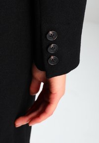 Musta villakangastakin hihassa on kolme pyöreää, tummaa nappia, joissa on neljä reikää. Pinnasta on pehmeä ja sileä. Käsi on lähellä.