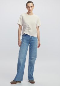 Beżowa koszulka z krótkim rękawem z haftem kwiatowym po lewej stronie, zestawiona z jasnoniebieskimi jeansami prostymi i szarym obuwiem.