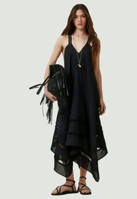 Vestido maxi preto sem mangas com decote em V, detalhes em recortes e bainha em camadas, combinado com uma mala preta com franjas e sandálias.