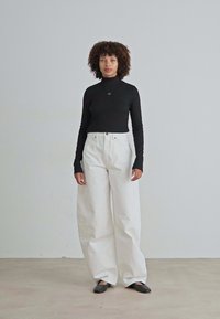 Zwarte lange mouwen turtleneck shirt gecombineerd met witte wijde broeken en zwarte flats, geplaatst tegen een neutrale achtergrond.