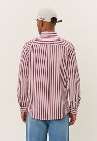 Cămașă cu dungi roșii și albe, cu guler ascuțit, mâneci lungi și margine rotundă. Asortată cu pantaloni din denim albastru deschis și o pălărie.