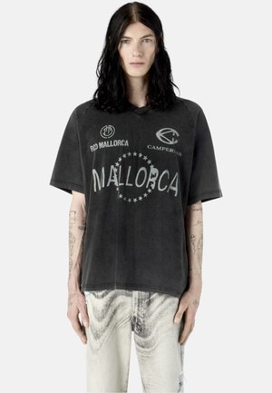 Person med langt sort hår bærer en falmet sort Mallorca grafisk t-shirt og lyse, mønstrede bukser, stående mod en ensfarvet baggrund.