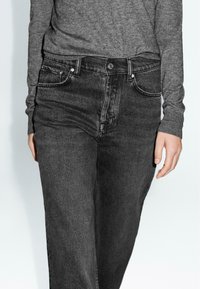 Jeans in denim grigio scuro a vita alta, con tasche frontali, passanti per cintura e chiusura a bottone. Texture liscia con sottili dettagli di sbiadimento.