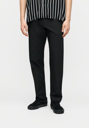 Άνδρας που φοράει ίσια μαύρα παντελόνια, μαύρα αθλητικά παπούτσια και μια μαύρη και άσπρη πουκάμισα με κάθετες ρίγες, στέκεται μπροστά σε λευκό φόντο.