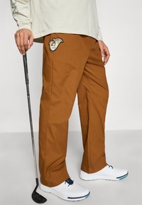 Pantalon marron de coupe décontractée avec un motif côté en patch. Porté avec des chaussures blanches, complété par un club de golf tenu dans la main droite.