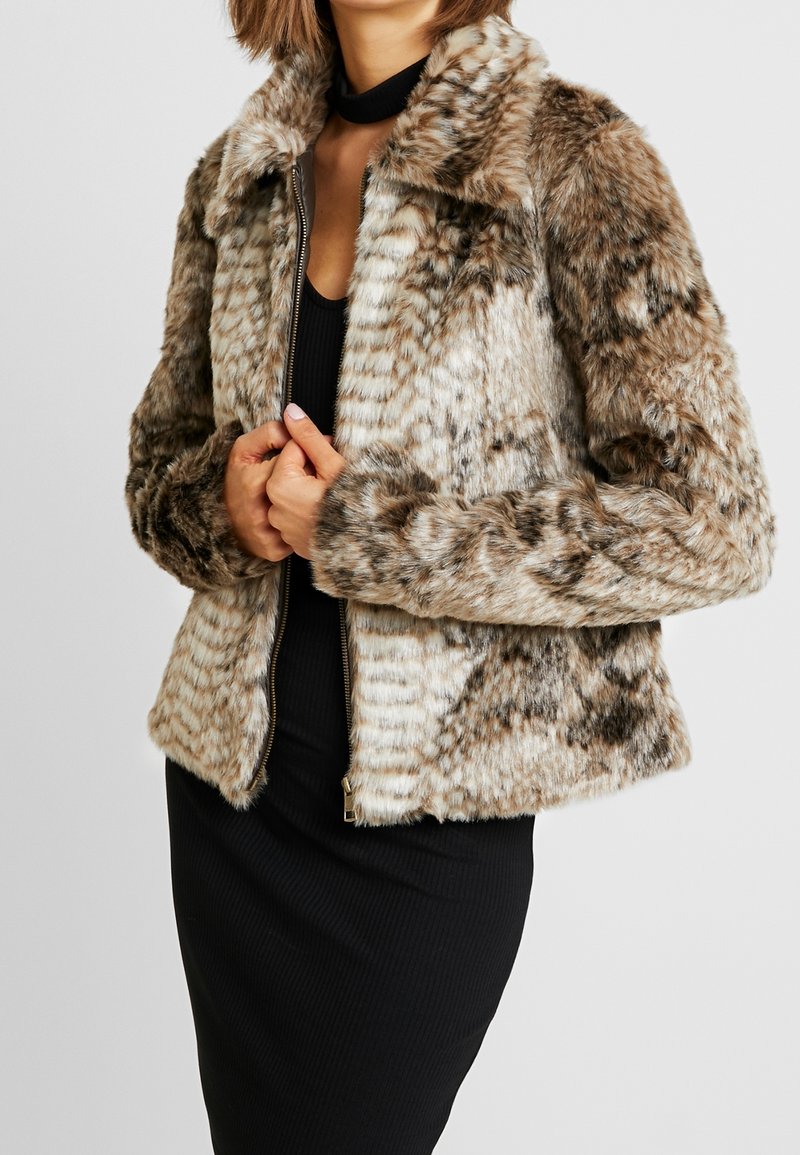 Femme portant une veste en fausse fourrure à motifs et zippée par-dessus une robe noire avec un col ras du cou, les mains près de la fermeture éclair.