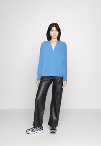 Maglietta blu a maniche lunghe con scollatura a V e polsini arricciati, abbinata a pantaloni di pelle nera e sneakers blu e argento. Tessuto liscio.