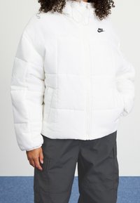 Vit vadderad jacka med hög krage, framdragkedja och svart Nike-logotyp på vänster sida. Designad med elastiska manchetter och sidofickor.