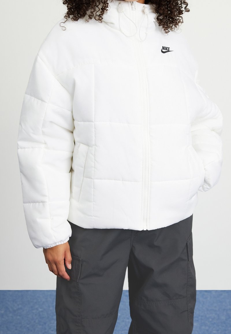 Vit vadderad jacka med hög krage, framdragkedja och svart Nike-logotyp på vänster sida. Designad med elastiska manchetter och sidofickor.