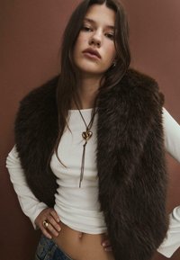 Hnědá kožešinová vesta přetažená přes bílý žebrovaný top. Modelka nosí zlatý náhrdelník ve tvaru srdce a prsten. Pozadí je v teplých tónech.