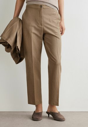 Person iført højtaljede brune, skræddersyede bukser og brune slip-on kitten heel-sko, der holder en matchende brun blazer i venstre hånd.