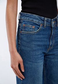 Dark blue denim jeans featuring a high waist, copper rivets, and subtle stitching details. The fabric has a smooth texture.