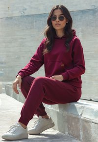 Burgundy kapucnis pulcsi elülső zsebel, hozzá illő melegítőnadrággal és fehér tornacipővel párosítva. Puha anyag, laza fazon, bordázott mandzsetta és alsó szegély.