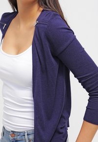 Cárdigan azul marino de manga larga, hecho de tela suave. Presenta herrajes plateados en los hombros y un diseño relajado y abierto.