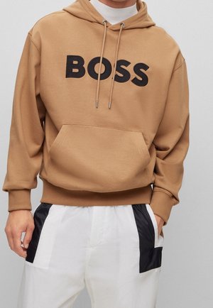 Sweat à capuche marron clair avec inscription noire « BOSS », poche avant, cordons, porté avec un pantalon blanc avec des panneaux latéraux noirs.
