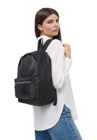 Zaino in tessuto nero con superficie testurizzata, tasca frontale con zip e spallacci regolabili, che si abbina a una camicia bianca e a jeans di denim.