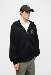 Nuori mies, jolla on päässään raidoitettu baseball-lippalakki, musta vetoketjulla varustettu huppari valkoisella logolla, valkoinen kauluspaita ja vihreät housut, seisoo kädet taskuissa.