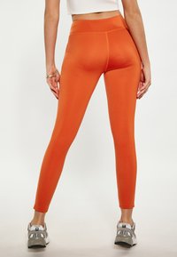 Oranje legging met hoge tailleband, gladde textuur en aansluitend ontwerp. Gecombineerd met grijze sportschoenen.