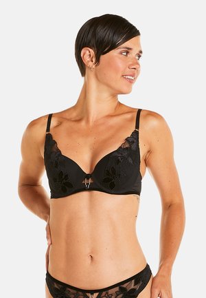 Soutien-gorge en dentelle noire avec broderie florale, bonnets opaques, bretelles ajustables et un accent décoratif central en métal.