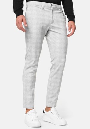 Homme portant un pantalon slim gris clair et des baskets blanches, debout les mains détendues le long du corps devant un fond uni.