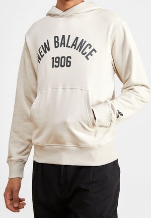 Beżowa bluza z kapturem z napisem "NEW BALANCE 1906" na przodzie, kieszenią kangurka i małym logo na lewym rękawie, noszona przez osobę w czarnych spodniach.