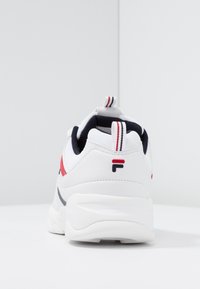 Baskets blanches à semelle épaisse, avec des accents rouges et bleus marine, des languettes à rayures, et un logo stylisé en forme de "F" sur le talon.