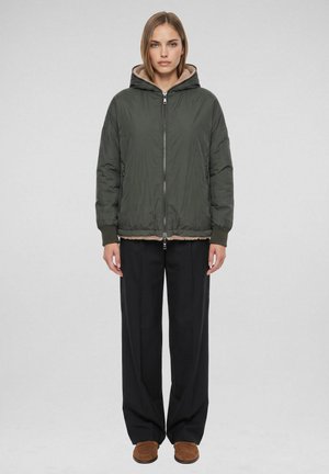 Vrouw staand, kijkend naar voren, draagt een donkergroene hoodie, zwarte wijde broekspijpen en bruine instapschoenen op een effen achtergrond.