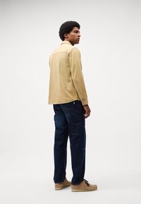 Jachetă bej din cord cu guler cu nasturi, purtată cu pantaloni cargo negri și pantofi din suede bej. Modelul stă cu fața întoarsă.