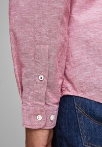 Camicia rossa intrecciata con maniche lunghe e polsini con bottone. Presenta una texture liscia, cuciture a contrasto e due bottoni bianchi sul polsino.