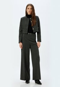 Dunkelgraue, kurz geschnittene Jacke mit goldenen Knöpfen, kombiniert mit weit geschnittenen grauen Hosen. Weiche Textur und schlanke Silhouette. Schwarzer Rollkragenpullover darunter.