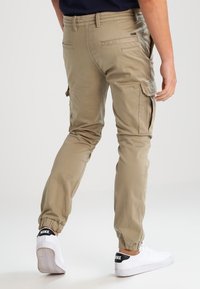 Kaki cargobyxor med elastiska mudd, flera sidoskickor och sömdetaljer. Hittas tillsammans med vita sneakers med svarta accenter.