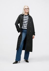Svart trenchcoat med dubbelknäppning, kombinerad med en randig skjorta och vida blå jeans. Svarta stövlar med klack fullbordar outfiten.