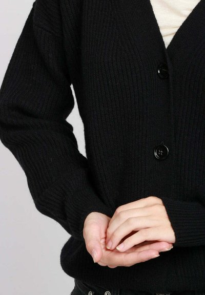 Persona che indossa un cardigan nero a coste abbottonato, con le mani delicatamente intrecciate davanti su uno sfondo chiaro e uniforme.