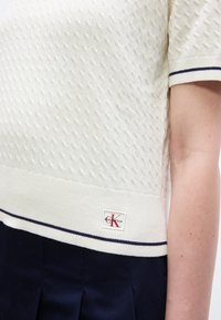 Λευκό πουλόβερ με κοντά μανίκια και υφή, με μαύρη επένδυση στα μανίκια και το τελείωμα, που φέρει ένα μικρό λογότυπο Calvin Klein πάνω από φούστα σε ναυτικό μπλε με πιέτες.