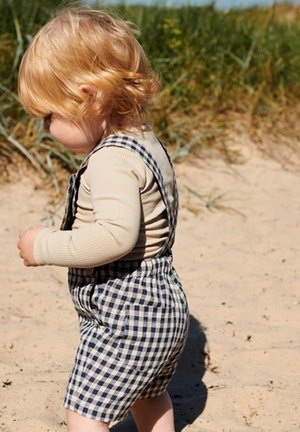 Toddler with blonde hair wearing beige long sleeve and blue checkered overalls walking on sandy beach with grass in the background.