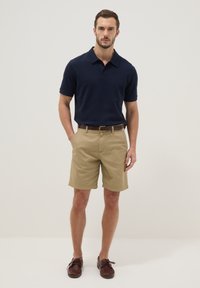 Hombre con polo azul marino, pantalones cortos beige, cinturón marrón y zapatos marrones con cordones, de pie contra un fondo claro y liso.