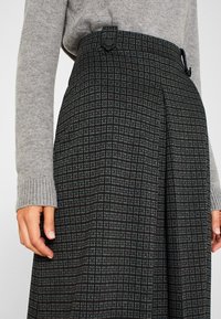 Jupe plissée en tissu foncé avec un motif à carreaux dans des tons de bleu et de vert. Taille haute avec fermeture à bouton et poches latérales.