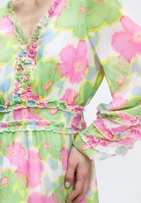 Κοντινό πλάνο παστέλ φλοράλ φόρεμα με βολάν στις άκρες και λεπτομέρειες με χρυσή κλωστή, που δείχνει μακριά φουσκωτά μανίκια και χέρι που ακουμπάει στη μέση.