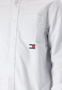 Camisa de botones en azul claro con finas rayas blancas, que cuenta con un bolsillo en el pecho y un pequeño logo de Tommy Jeans. Tejido de algodón, diseño casual.