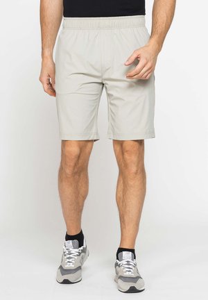 Uomo che indossa pantaloncini beige a vita elastica e scarpe da corsa grigie con calzini neri alla caviglia, in piedi contro uno sfondo bianco.