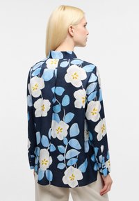 Blomstret langærmet skjorte i marineblå med store hvide og lyseblå blomster, gule accenter og en afslappet pasform. Knaplukning vedærmerne og detalje med rygfold.