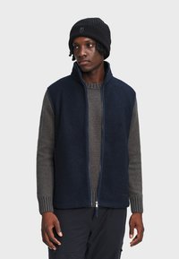 Gilet in pile blu navy con collo alto, zip completa e tasche laterali, indossato sopra un maglione grigio in maglia. Accessorio: berretto nero a coste.