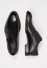 Chaussures en cuir noir avec un motif en chevrons, dotées d'une fermeture éclair sur le côté et d'un talon rectangulaire. L'intérieur affiche "HUGO" en rouge.