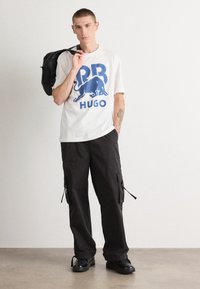 Hvid T-shirt med blåt grafisk design, der forestiller en tyr og teksten "HUGO". Mørke cargo bukser med sidelommer og sorte sko. Sort rygsæk.