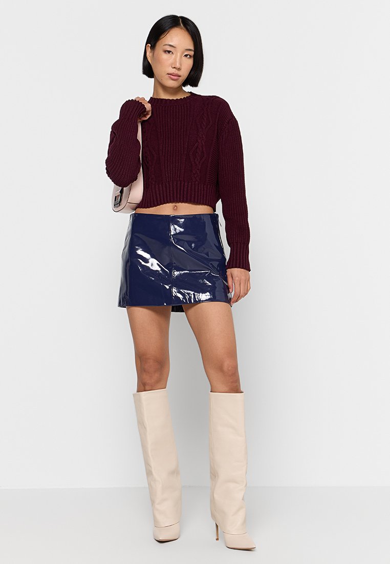 Maglione corto a maglia color bordeaux, minigonna lucida in patent blu navy, stivali al ginocchio beige e borsa rosa chiaro. Texture morbide e design aderente.