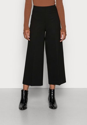 Pantalons noirs à larges jambes et taille haute, fabriqués en tissu lisse. Portés avec des bottines noires à motif crocodile.