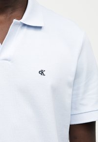 Άνδρας που φοράει γαλάζιο πόλο με μικρό μαύρο λογότυπο "cK" στο αριστερό στήθος σε μονόχρωμο φόντο.