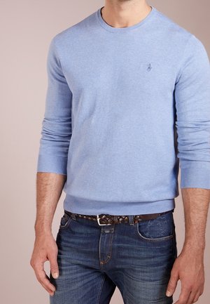 Hombre con suéter de manga larga azul claro con un pequeño logo en el pecho y vaqueros azul oscuro con cinturón trenzado marrón.