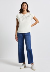 Light-colored short-sleeve top with "Very Easy" printed in script; worn with blue wide-leg jeans and white sneakers, featuring a relaxed fit.